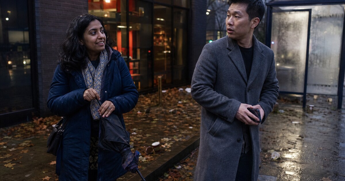 A 32-year-old South Asian woman in a worn navy puffer coat and a 35-year-old East Asian man in a plain grey wool coat greet each other a…