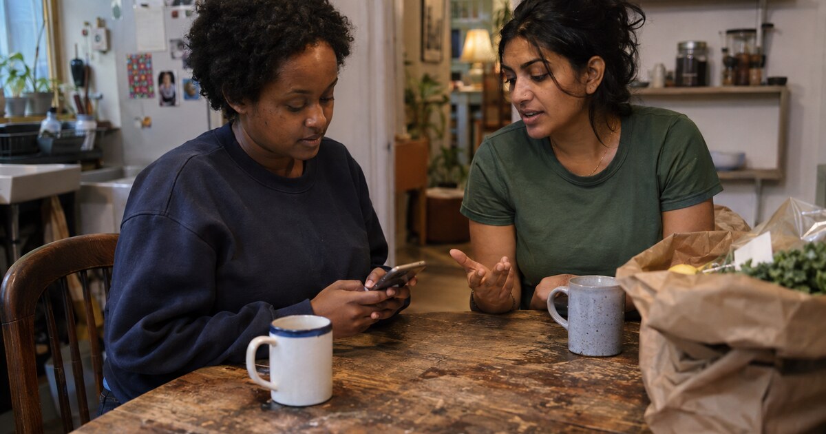 A Black woman in her early thirties with short natural hair and a faded navy sweatshirt types a message on her phone with both thumbs at a…