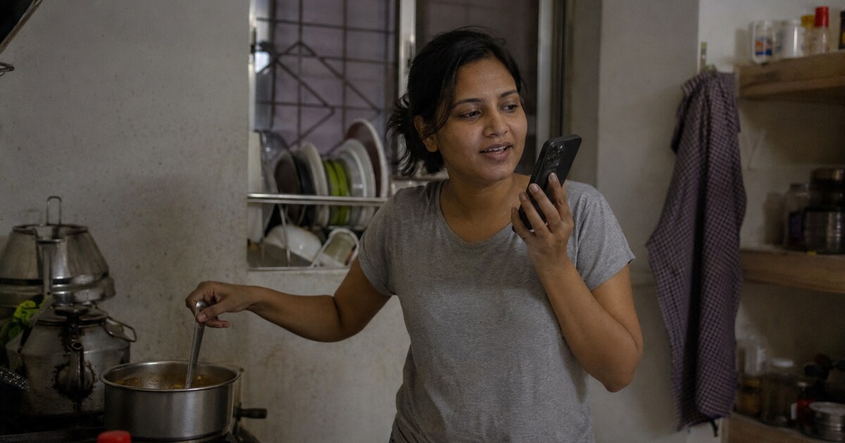 A South Asian woman in her early thirties with loosely tied dark hair, wearing a plain grey T-shirt and checked pyjama trousers, stands in…